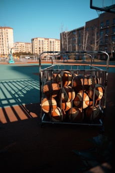 A cart filled with basketballs is positioned on an outdoor basketball court. The court is illuminated by sunlight, creating shadows and a vibrant atmosphere. In the background, several residential buildings surround the area, adding an urban feel to the setting.