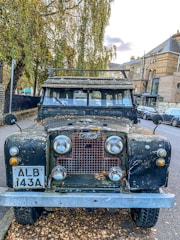 An old Land Rover parked on the side of a street, showing signs of wear with rust and paint peeling off. It is surrounded by autumn leaves and there are trees with fall foliage nearby. The background features brick buildings and a few parked cars.