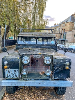 An old Land Rover parked on the side of a street, showing signs of wear with rust and paint peeling off. It is surrounded by autumn leaves and there are trees with fall foliage nearby. The background features brick buildings and a few parked cars.