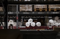 A warehouse shelf filled with packaged rolls of material. The rolls are arranged neatly on metal racks, surrounded by boxes and pallets. The lighting is dim, creating a sense of depth and storage abundance.