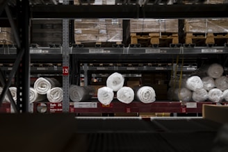 A warehouse shelf filled with packaged rolls of material. The rolls are arranged neatly on metal racks, surrounded by boxes and pallets. The lighting is dim, creating a sense of depth and storage abundance.