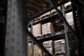 Large pallets wrapped in plastic are stacked on several levels of metal shelving inside a warehouse. The focus is on the orderly arrangement of goods, emphasizing industrial efficiency.