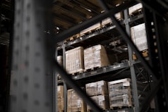 Large pallets wrapped in plastic are stacked on several levels of metal shelving inside a warehouse. The focus is on the orderly arrangement of goods, emphasizing industrial efficiency.