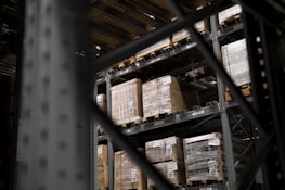 Large pallets wrapped in plastic are stacked on several levels of metal shelving inside a warehouse. The focus is on the orderly arrangement of goods, emphasizing industrial efficiency.