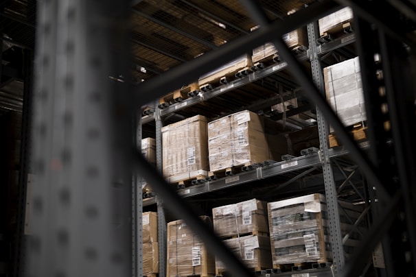 Large pallets wrapped in plastic are stacked on several levels of metal shelving inside a warehouse. The focus is on the orderly arrangement of goods, emphasizing industrial efficiency.