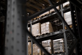 Large pallets wrapped in plastic are stacked on several levels of metal shelving inside a warehouse. The focus is on the orderly arrangement of goods, emphasizing industrial efficiency.