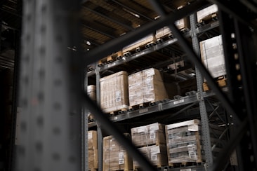 Large pallets wrapped in plastic are stacked on several levels of metal shelving inside a warehouse. The focus is on the orderly arrangement of goods, emphasizing industrial efficiency.