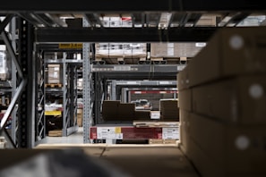 A warehouse interior with rows of metal shelving units filled with cardboard boxes and pallets. The shelves extend into the distance, creating a symmetrical, repetitive pattern. Overhead signs indicate row numbers and height restrictions. The lighting is dim and industrial.