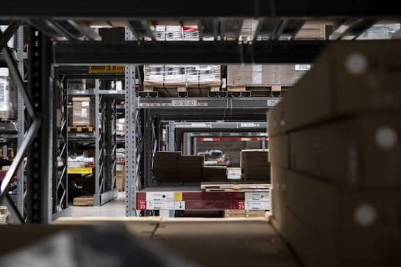 A warehouse interior with rows of metal shelving units filled with cardboard boxes and pallets. The shelves extend into the distance, creating a symmetrical, repetitive pattern. Overhead signs indicate row numbers and height restrictions. The lighting is dim and industrial.