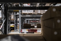 A warehouse interior with rows of metal shelving units filled with cardboard boxes and pallets. The shelves extend into the distance, creating a symmetrical, repetitive pattern. Overhead signs indicate row numbers and height restrictions. The lighting is dim and industrial.