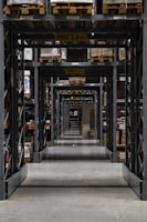 Long warehouse aisle with tall metal shelving units stacked with wooden pallets. The perspective creates a series of repetitive frames that lead the eye to the back of the warehouse. The concrete floor is clean and polished, while the shelves are organized and labeled. Ambient lighting casts soft shadows.