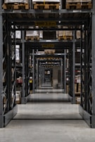 Long warehouse aisle with tall metal shelving units stacked with wooden pallets. The perspective creates a series of repetitive frames that lead the eye to the back of the warehouse. The concrete floor is clean and polished, while the shelves are organized and labeled. Ambient lighting casts soft shadows.