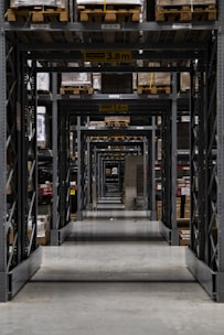 Long warehouse aisle with tall metal shelving units stacked with wooden pallets. The perspective creates a series of repetitive frames that lead the eye to the back of the warehouse. The concrete floor is clean and polished, while the shelves are organized and labeled. Ambient lighting casts soft shadows.