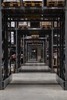 Long warehouse aisle with tall metal shelving units stacked with wooden pallets. The perspective creates a series of repetitive frames that lead the eye to the back of the warehouse. The concrete floor is clean and polished, while the shelves are organized and labeled. Ambient lighting casts soft shadows.