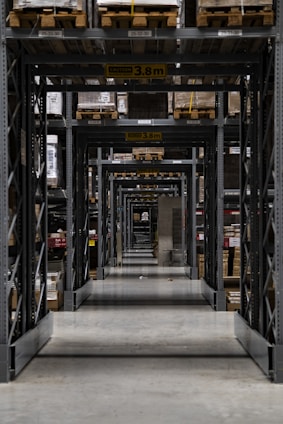 Long warehouse aisle with tall metal shelving units stacked with wooden pallets. The perspective creates a series of repetitive frames that lead the eye to the back of the warehouse. The concrete floor is clean and polished, while the shelves are organized and labeled. Ambient lighting casts soft shadows.
