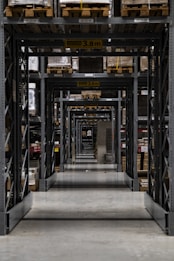 Long warehouse aisle with tall metal shelving units stacked with wooden pallets. The perspective creates a series of repetitive frames that lead the eye to the back of the warehouse. The concrete floor is clean and polished, while the shelves are organized and labeled. Ambient lighting casts soft shadows.