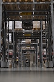 A large warehouse aisle is filled with tall shelves stacked with pallets and boxes. The perspective creates a tunnel-like view with overhead caution signs indicating maximum height clearance. Rows of metal beams and industrial shelving dominate the space, with the floor appearing smooth and reflective.