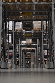 A large warehouse aisle is filled with tall shelves stacked with pallets and boxes. The perspective creates a tunnel-like view with overhead caution signs indicating maximum height clearance. Rows of metal beams and industrial shelving dominate the space, with the floor appearing smooth and reflective.