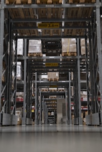 A large warehouse aisle is filled with tall shelves stacked with pallets and boxes. The perspective creates a tunnel-like view with overhead caution signs indicating maximum height clearance. Rows of metal beams and industrial shelving dominate the space, with the floor appearing smooth and reflective.