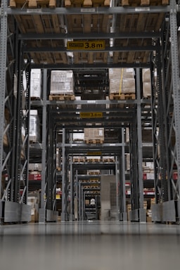 A large warehouse aisle is filled with tall shelves stacked with pallets and boxes. The perspective creates a tunnel-like view with overhead caution signs indicating maximum height clearance. Rows of metal beams and industrial shelving dominate the space, with the floor appearing smooth and reflective.
