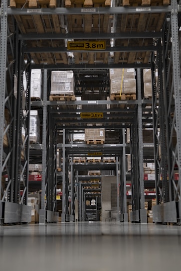 A large warehouse aisle is filled with tall shelves stacked with pallets and boxes. The perspective creates a tunnel-like view with overhead caution signs indicating maximum height clearance. Rows of metal beams and industrial shelving dominate the space, with the floor appearing smooth and reflective.