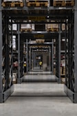 A long corridor in a warehouse features tall industrial metal shelving units filled with various boxed and palletized goods. At the end of the corridor, a person in a yellow jacket is visible. The space is organized with caution signs indicating height restrictions.