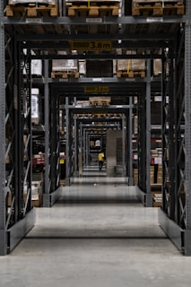 A long corridor in a warehouse features tall industrial metal shelving units filled with various boxed and palletized goods. At the end of the corridor, a person in a yellow jacket is visible. The space is organized with caution signs indicating height restrictions.