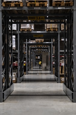 A long corridor in a warehouse features tall industrial metal shelving units filled with various boxed and palletized goods. At the end of the corridor, a person in a yellow jacket is visible. The space is organized with caution signs indicating height restrictions.