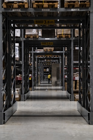 A long corridor in a warehouse features tall industrial metal shelving units filled with various boxed and palletized goods. At the end of the corridor, a person in a yellow jacket is visible. The space is organized with caution signs indicating height restrictions.