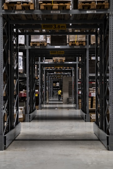 A long corridor in a warehouse features tall industrial metal shelving units filled with various boxed and palletized goods. At the end of the corridor, a person in a yellow jacket is visible. The space is organized with caution signs indicating height restrictions.