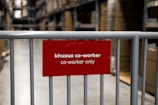 A red sign attached to a metal barrier indicates a restricted area for coworkers only. The background features shelves and boxes, suggesting a warehouse or storage environment.