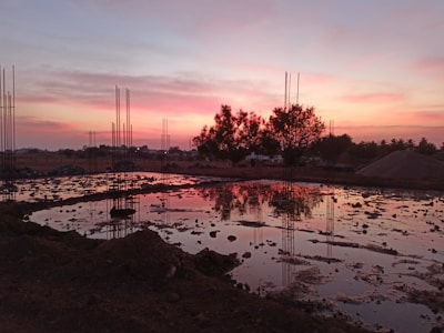 A construction site is depicted at sunset with exposed rebar rods protruding from the ground. The sky is a blend of pink and orange hues, reflecting on the water-filled excavation. Trees are silhouetted against the colorful sky, and piles of dirt are visible.