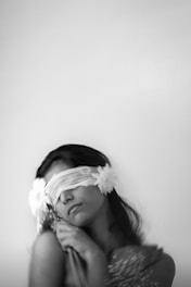 A blurred black-and-white photograph portrays a woman with a blindfold, adorned with flowers near her head. She appears to be in a state of introspection or contemplation, with her eyes covered and head slightly tilted.