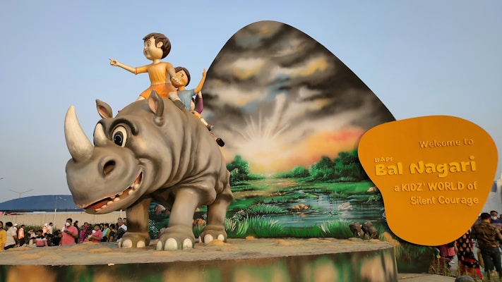 A large statue of a rhino with two children sitting on its back is central to the scene. The background features a painted landscape with a setting or rising sun. On the right, a yellow sign reads 'Welcome to BAPI Bal Nagari, a KIDZ' WORLD of Silent Courage'. A crowd of people is gathered around.