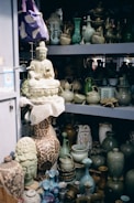A collection of ceramic and pottery items, including vases, teapots, and decorative figurines, displayed on shelves. A prominent large ceramic Buddha statue is also visible, sitting on a shelf amid various artfully crafted pottery pieces. The color palette is predominantly earthy with green and blue hues.