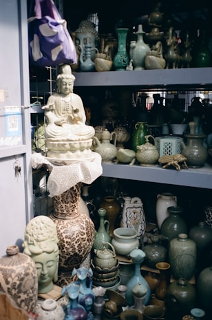 A collection of ceramic and pottery items, including vases, teapots, and decorative figurines, displayed on shelves. A prominent large ceramic Buddha statue is also visible, sitting on a shelf amid various artfully crafted pottery pieces. The color palette is predominantly earthy with green and blue hues.