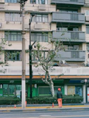 A person wearing an orange helmet and pants stands in front of a residential building with balconies. The building is light-colored with dark gray balconies and window frames. There are multiple air conditioning units attached to the building. A tree and some bushes are located near the sidewalk. Traffic cones are visible on the road.