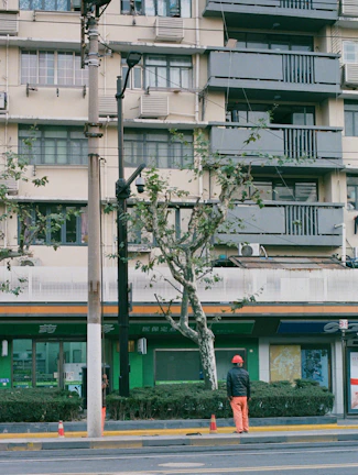 A person wearing an orange helmet and pants stands in front of a residential building with balconies. The building is light-colored with dark gray balconies and window frames. There are multiple air conditioning units attached to the building. A tree and some bushes are located near the sidewalk. Traffic cones are visible on the road.