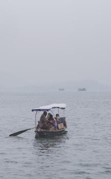 A small wooden boat is on calm, grey water, with a family of three on board. The boat is covered with a simple canopy, and each person is warmly dressed. The background is misty, suggesting a foggy or overcast day with distant and barely visible larger boats.