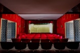 A small, intimate theater with red velvet seats arranged in rows facing a stage with a closed curtain. The walls are covered in matching red velvet paneling, and there are decorative lamps on the side walls. The stage is framed with a red curtain and light-colored backdrop.