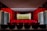A small, intimate theater with red velvet seats arranged in rows facing a stage with a closed curtain. The walls are covered in matching red velvet paneling, and there are decorative lamps on the side walls. The stage is framed with a red curtain and light-colored backdrop.