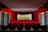 A small, intimate theater with red velvet seats arranged in rows facing a stage with a closed curtain. The walls are covered in matching red velvet paneling, and there are decorative lamps on the side walls. The stage is framed with a red curtain and light-colored backdrop.