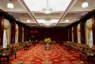 A luxurious and formal room with an ornate red carpet covering the floor. Above, multiple chandeliers hang from the ceiling, enhancing the room's elegance. On either side, cushioned sofas line the walls, accompanied by dark wooden coffee tables. At the far end, there are flags on display and a podium adorned with floral arrangements.