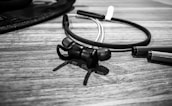 A pair of wireless earphones placed on a wooden surface. The earphones have a sleek design with in-ear tips, connected by a flexible band. Nearby, there are pen caps or similar objects that suggest a casual and creative environment.