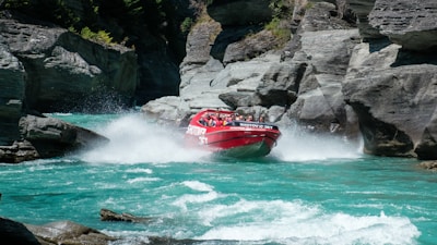 A red jet boat navigates through a narrow gorge with turquoise water, surrounded by steep, rocky cliffs. Water splashes dramatically as the boat speeds through the river, and passengers are visible on board experiencing the thrill.