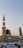 A beautiful Islamic mosque features a tall minaret adorned with green lighting at the top, complemented by a striking green dome below. Underneath, a gathering of people in white traditional attire is observable, suggesting a spiritual or communal setting during dawn or dusk.