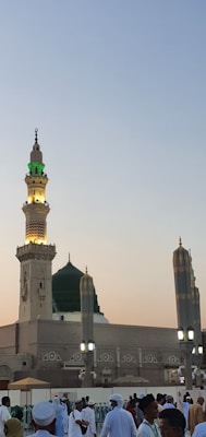 A beautiful Islamic mosque features a tall minaret adorned with green lighting at the top, complemented by a striking green dome below. Underneath, a gathering of people in white traditional attire is observable, suggesting a spiritual or communal setting during dawn or dusk.