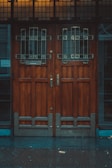 A pair of large wooden doors with intricate metal grillwork and stained glass panels at the top. The wood appears dark and polished, adding to the vintage aesthetic. The base of the doors includes metal reinforcement. The area surrounding the doors is dimly lit, suggesting an overcast day or evening.