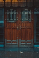 A pair of large wooden doors with intricate metal grillwork and stained glass panels at the top. The wood appears dark and polished, adding to the vintage aesthetic. The base of the doors includes metal reinforcement. The area surrounding the doors is dimly lit, suggesting an overcast day or evening.