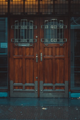 A pair of large wooden doors with intricate metal grillwork and stained glass panels at the top. The wood appears dark and polished, adding to the vintage aesthetic. The base of the doors includes metal reinforcement. The area surrounding the doors is dimly lit, suggesting an overcast day or evening.